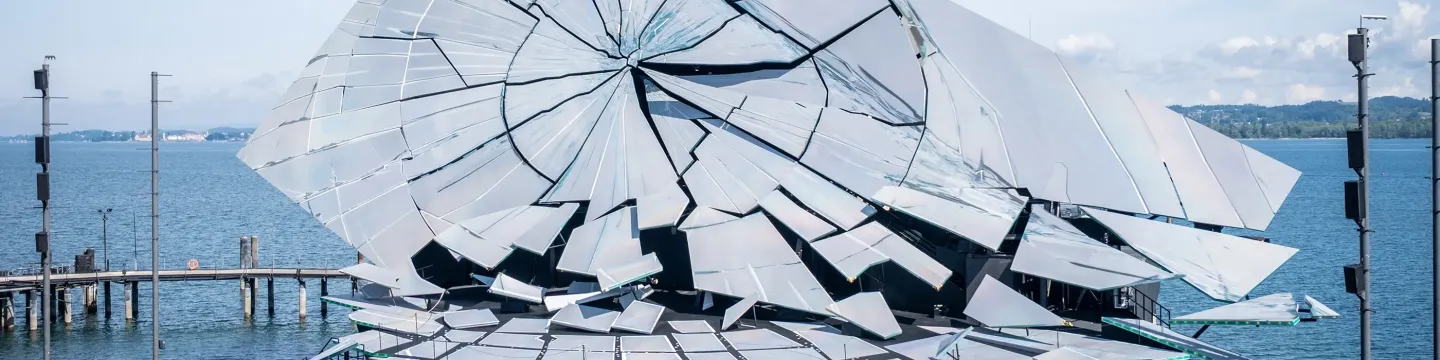 © Bregenzer Festspiele / Kristina Becker Große, zerbrochene Spiegelinstallation auf einem Wasserbecken mit Stegen und See im Hintergrund