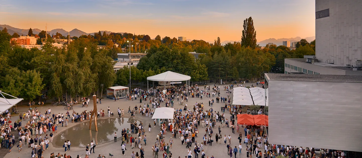 Großer Vorplatz vor dem Festspielhaus Bregenz mit vielen Menschen bei Sonnenuntergang, umgeben von Bäumen und Bergen im Hintergrund