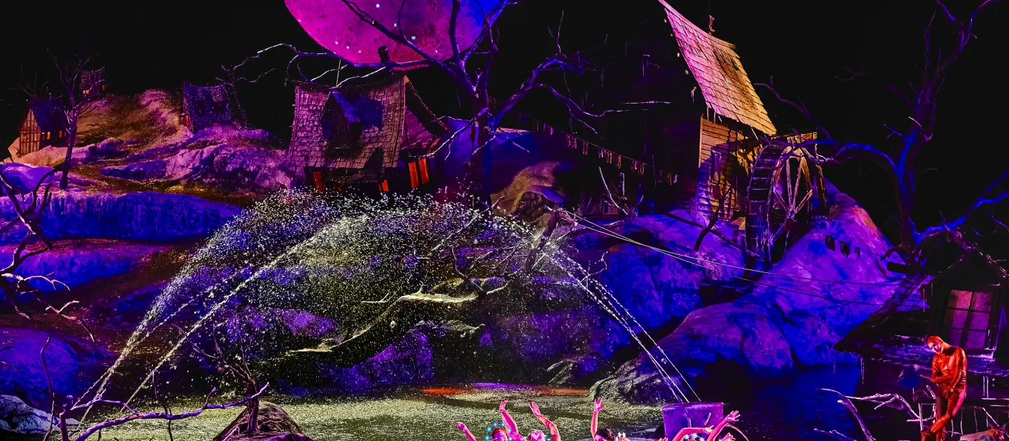 Bühnenbild von 'Der Freischütz' mit Tänzern in farbenfrohen Kostümen vor einem großen, leuchtenden Vollmond und einer mystischen Waldkulisse