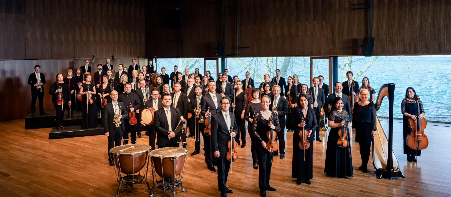 Symphonieorchester Vorarlberg mit Musikerinnen und Musikern in formeller Kleidung, stehend mit ihren Instrumenten in einem großen Konzertsaal mit Holzwänden und großen Fenstern