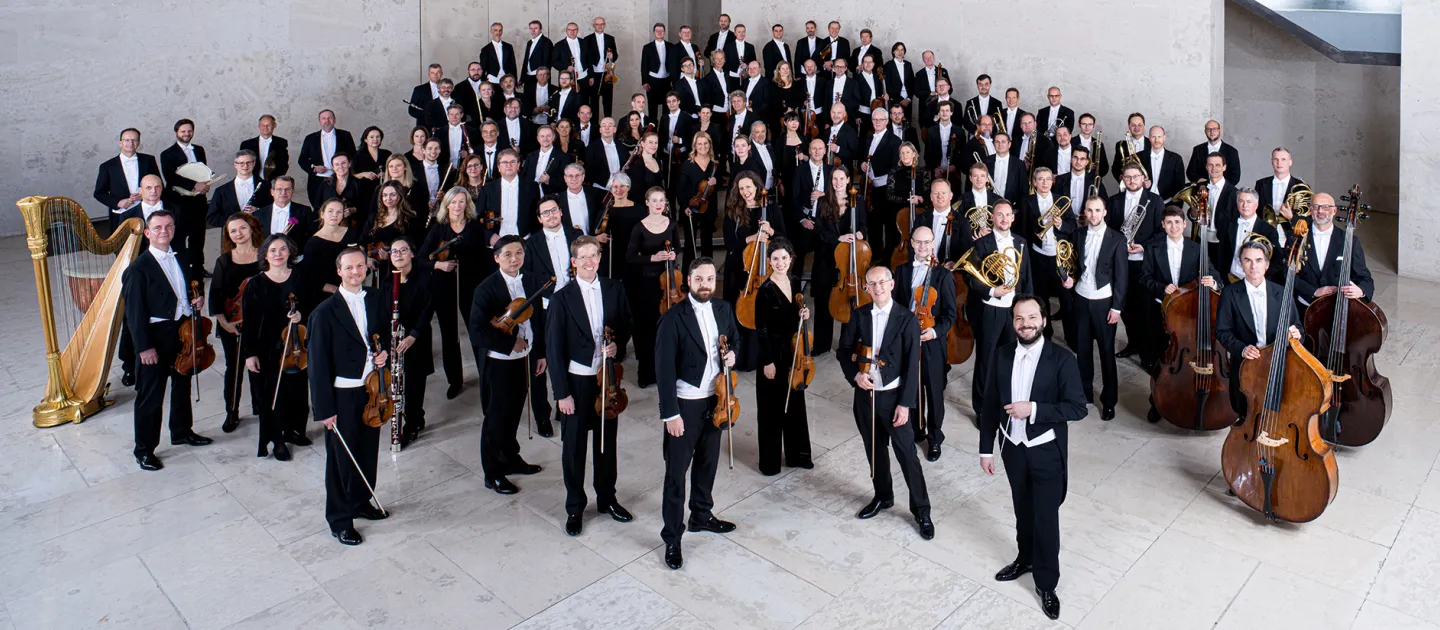 Mitglieder der Wiener Symphoniker in formeller Konzertkleidung mit Instrumenten, auf einer Treppe in einem modernen Saal gruppiert