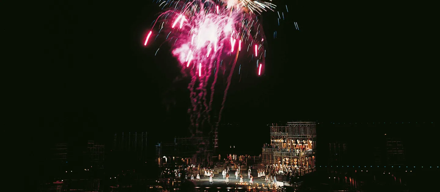 © Bregenzer Festspiele Szene aus "Eine Nacht in Venedig", 1975 auf der Seebühne, dreistöckiges Gebäude, viele Darstellende in Kostümen, am Himmel ist ein buntes Feuerwerk über dem Bühnenbild
