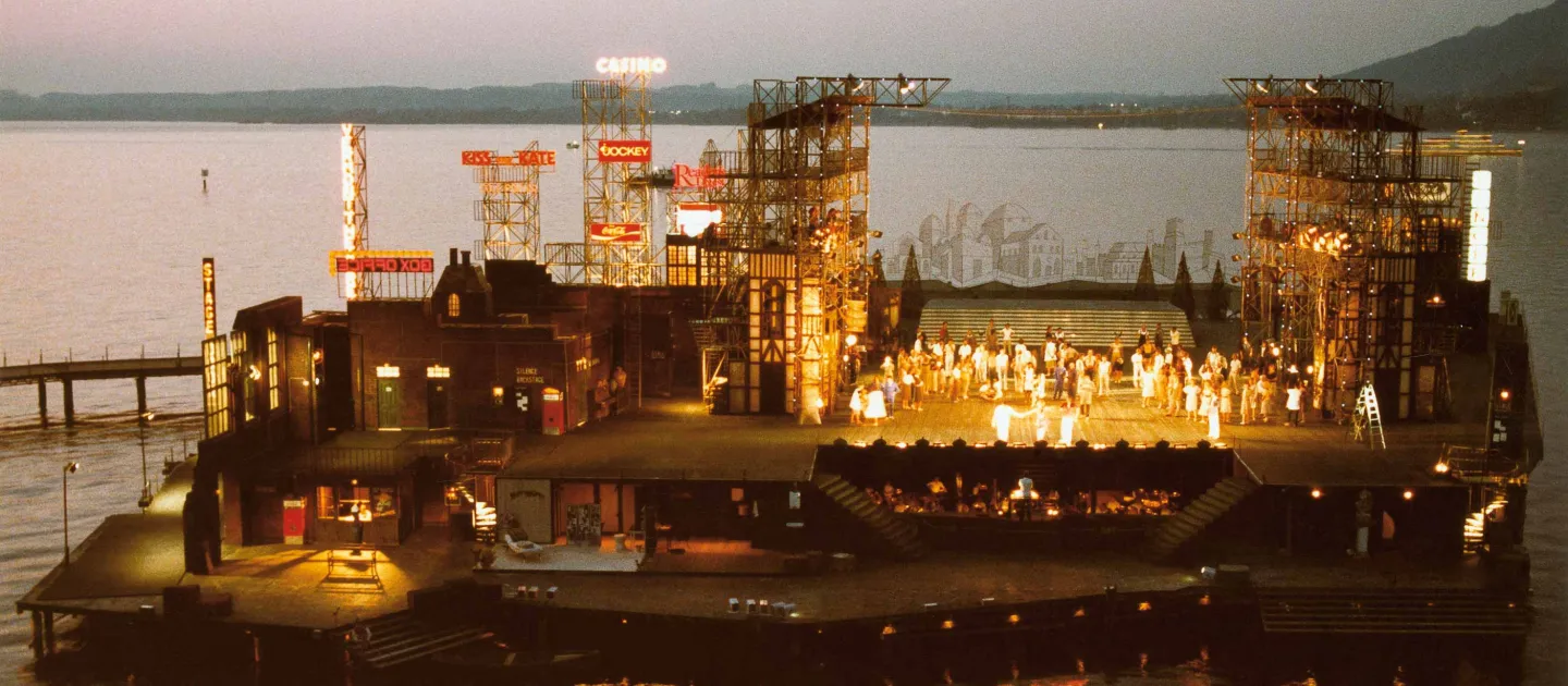 Szene aus "Kiss me, Kate", 1983 auf der Seebühne, zweistöckiges Bühnenbild, obere Ebene mit Gebäuden und industriellen Gerüstkonstruktionen, in der rechten Bühnenhälfte befinden sich kostümierte Darstellende