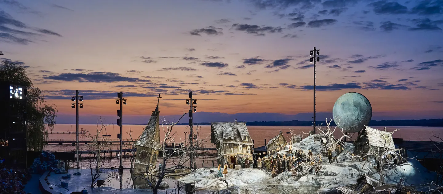 Bühnenbild 'Der Freischütz' mit unwirtlichem Dorf und Mond im Hintergrund, sowie einem Kirchturm. Hinter dem Bühnenbild geht im Wasser die Sonne unter