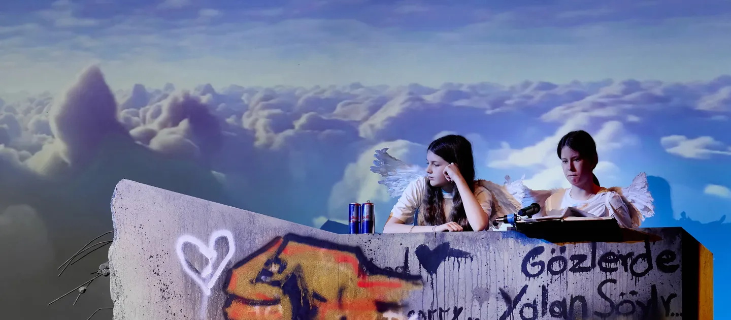 © Bregenzer Festspiele / Anja Koehler Zwei Personen mit Flügeln sitzen auf einer grauen Wand mit Graffiti. Die Wand zeigt eine große, bunte Hand und verschiedene Symbole wie Herzen und Frieden. Im Hintergrund sind Wolken zu sehen.