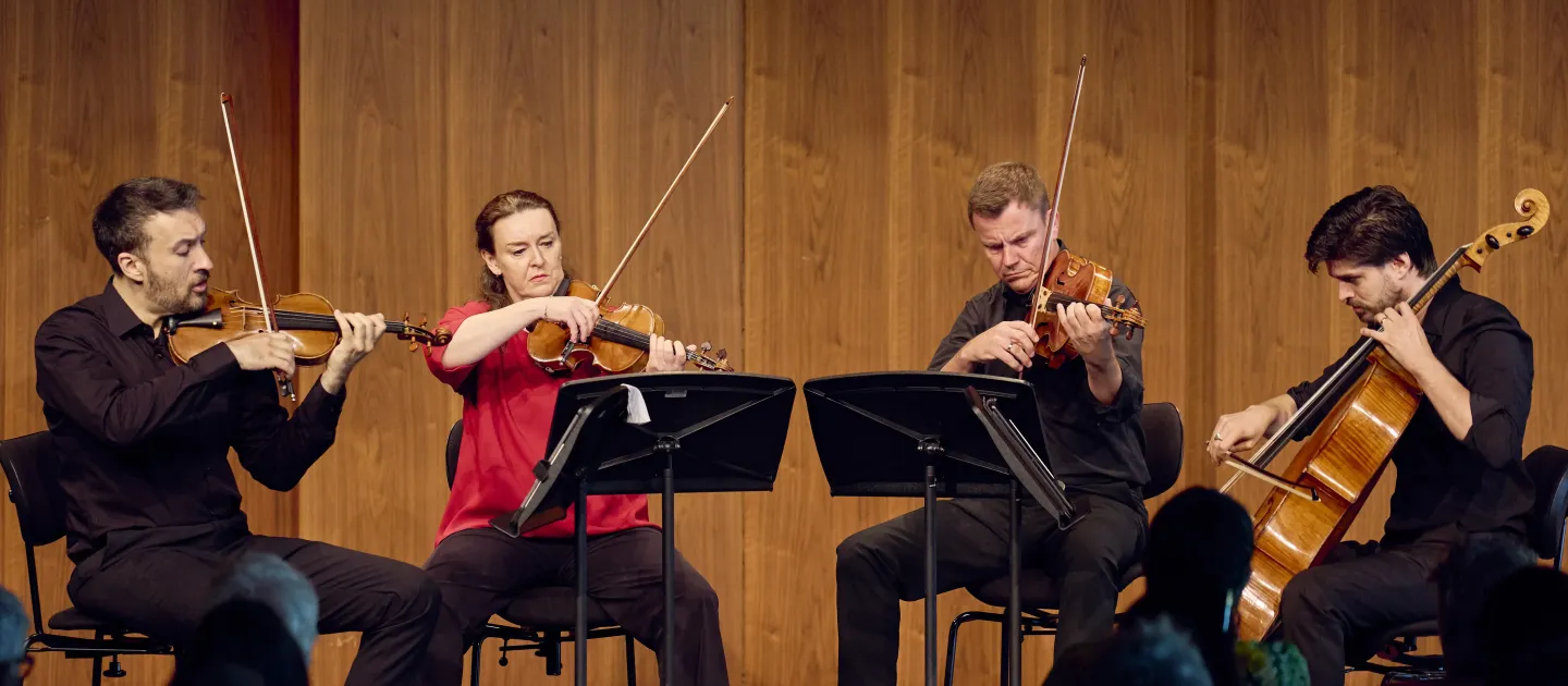 Vier Musiker in schwarzer Kleidung spielen ein Streichquartett auf der Bühne. Zwei Violinisten, ein Bratschist und ein Cellist stehen in einer Reihe hinter Notenständern.