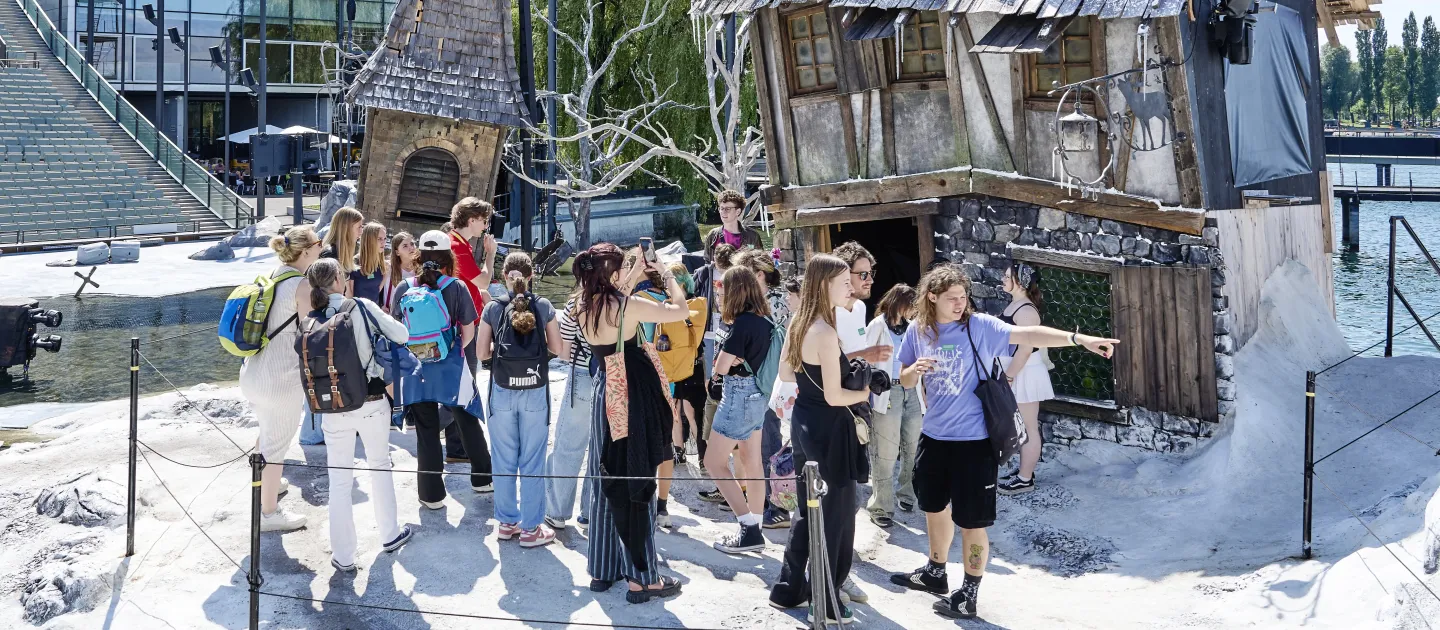 © Bregenzer Festspiele / Anja Koehler Gruppe junger Menschen steht vor einem großen, alten, schiefen Haus mit einem steinernen Kamin und einem Baum. Im Hintergrund sind moderne Gebäude und Lichtanlagen sichtbar.