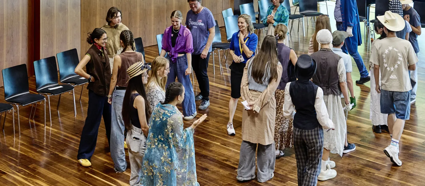 © Bregenzer Festspiele / Anja Koehler Eine Gruppe von jungen Menschen in verschiedenen Outfits interagiert in einem Raum mit Holzboden. Einige tragen Hüte und Accessoires, während andere in lässiger Kleidung gekleidet sind. Stühle stehen im Hintergrund.