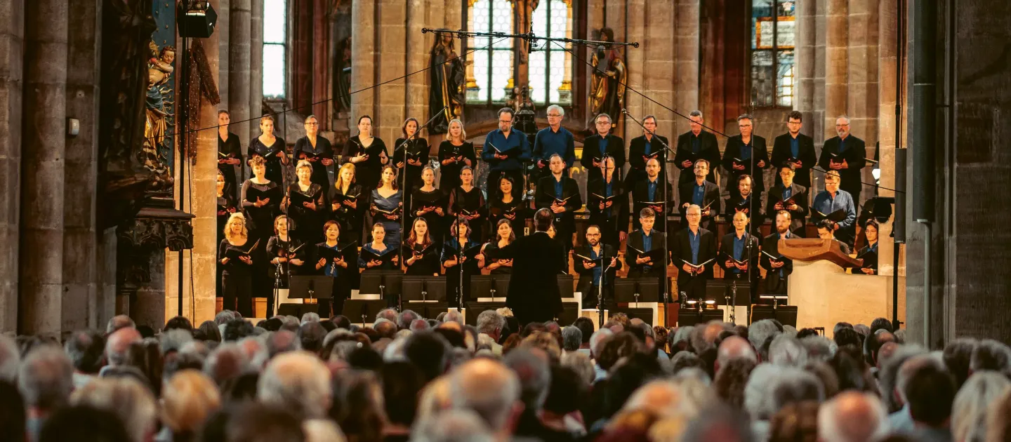 © Philip Kreibig Großer Chor mit mehreren Sängerinnen und Sängern in dunkler Kleidung steht auf Stufen in einer Kirche vor Publikum, Dirigent mit erhobenen Armen.