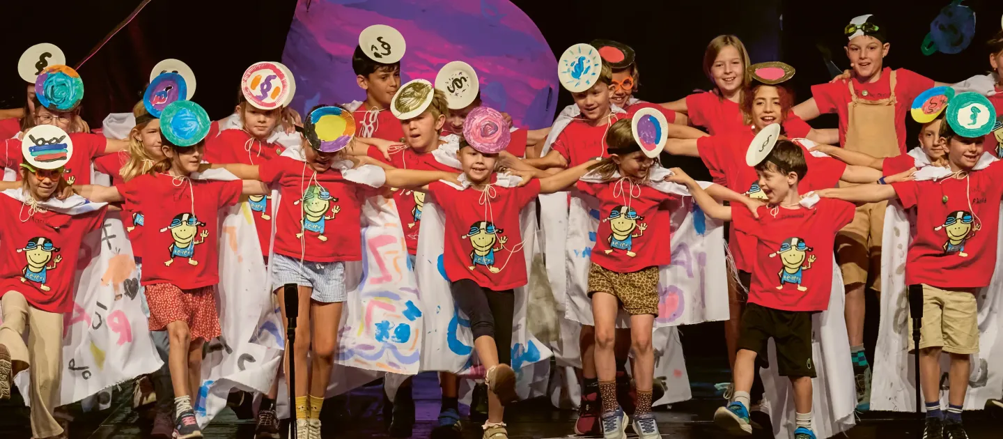 © Bregenzer Festspiele / Anja Koehler Gruppe von Kindern in roten T-Shirts mit Comicfigur, die auf einer Bühne mit buntem Hintergrund Arm in Arm stehen.