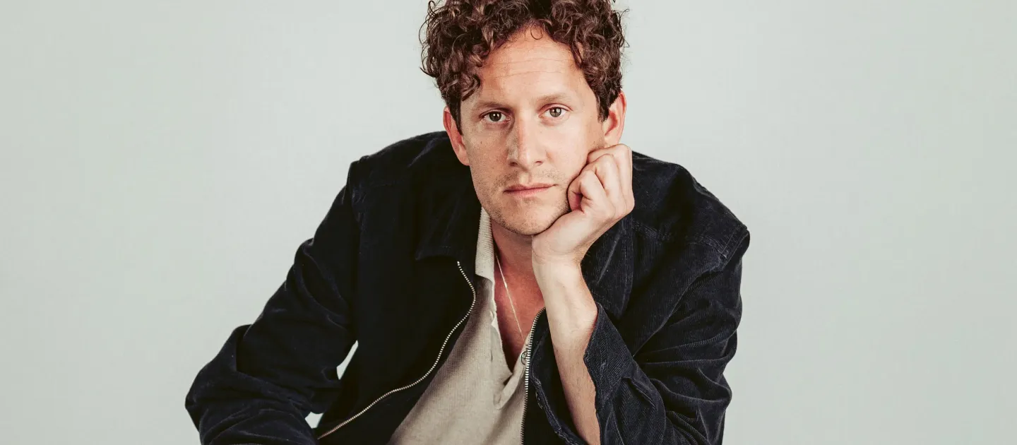 Junger Mann mit lockigem Haar sitzt mit gebeugtem Arm und stützt den Kopf auf die Hand, trägt helles Hemd und dunkle Jacke.