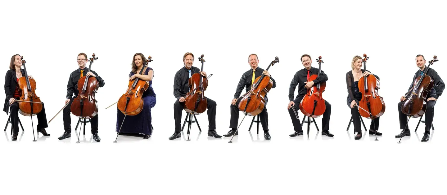 © Fotobyhofer Sieben Musiker sitzen in einer Reihe, jeder mit einem Cello, alle tragen schwarze Kleidung, vor weißem Hintergrund.