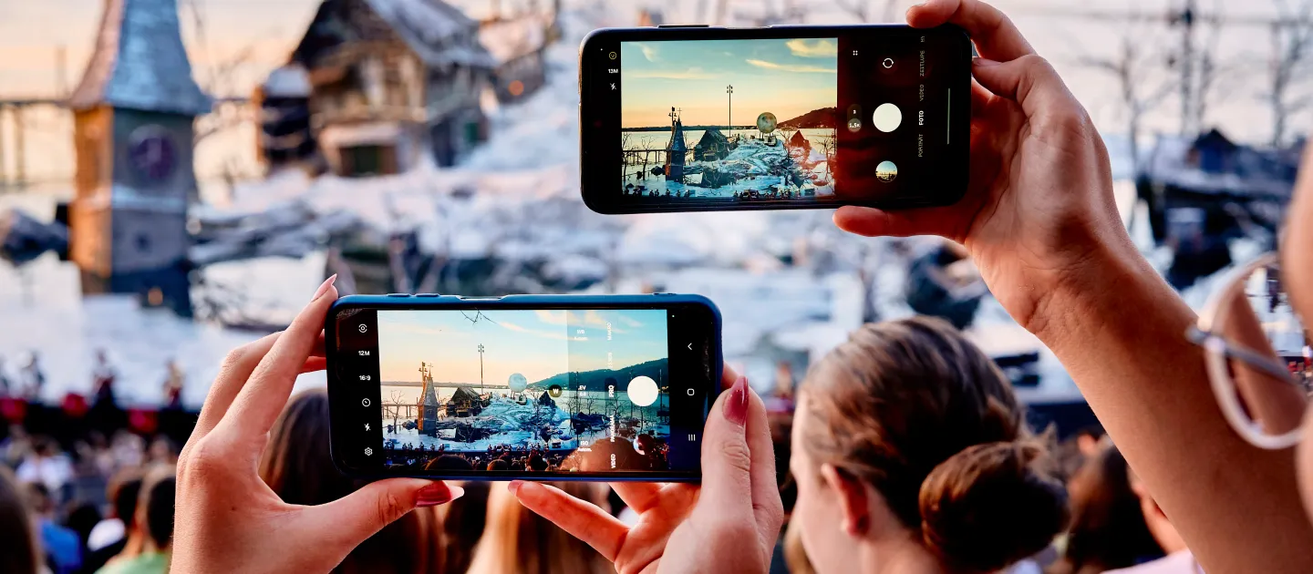 © Bregenzer Festspiele / Anja Koehler Zwei Hände halten Smartphones, die eine winterliche Stadtansicht mit Kirchturm und Bergen im Hintergrund fotografieren.