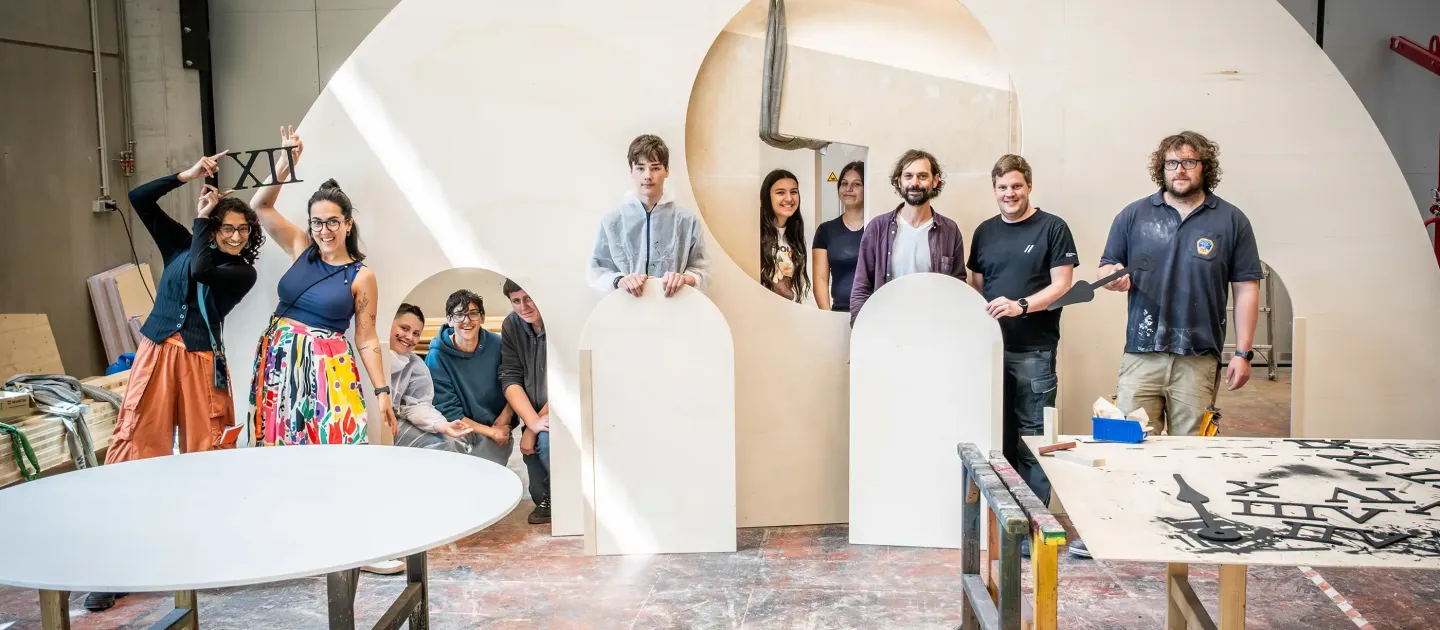 Gruppe von neun Personen steht und sitzt in einer Werkstatt hinter großen, weißen Buchstaben und Formen aus Holz