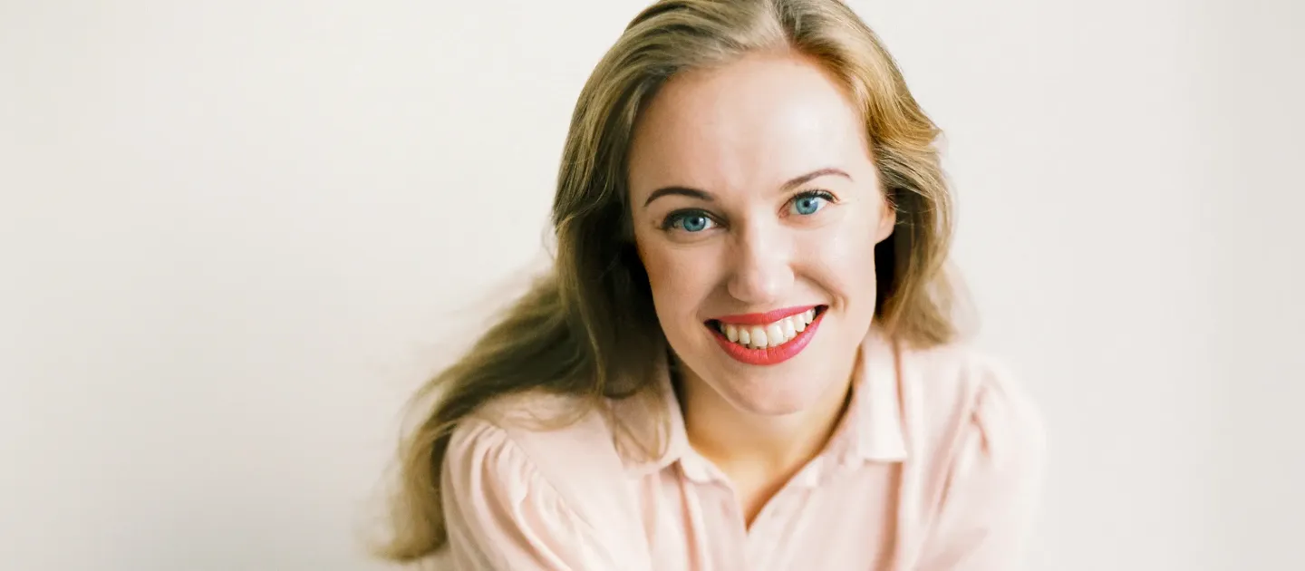 Lächelnde Frau mit langen blonden Haaren und hellrosa Bluse vor hellem Hintergrund