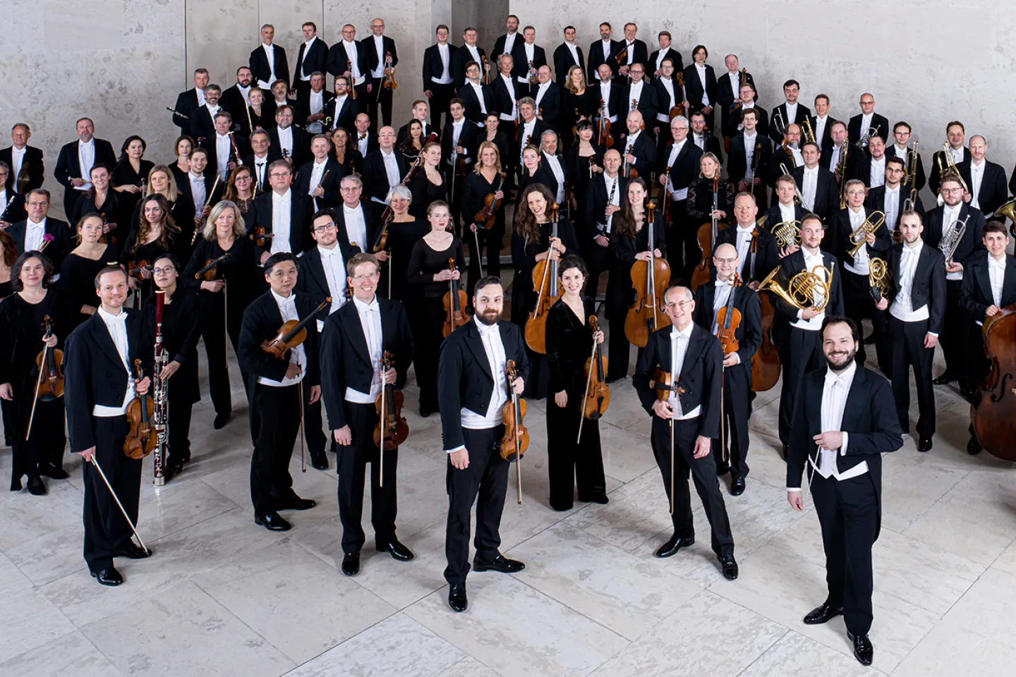 © Bregenzer Festspiele / Peter Rigaud Mitglieder der Wiener Symphoniker in formeller Konzertkleidung mit Instrumenten, auf einer Treppe in einem modernen Saal gruppiert