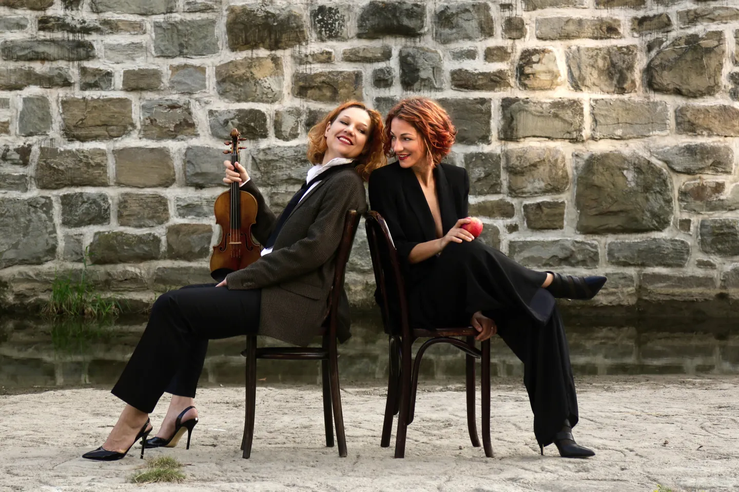 © Phoebe Violet Sophie Heinrich und Maria Radutu sitzen Rücken an Rücken auf Stühlen vor einer Steinmauer, Sophie hält eine Geige und beide lächeln entspannt