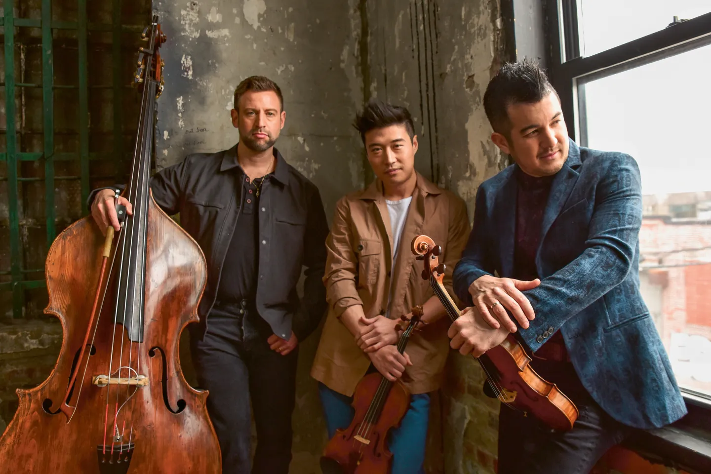 Musikalisches Trio time for Three mit drei Männern, einer hält einen Kontrabass, zwei halten Violinen, in einem Raum mit roher Wand und Fenster