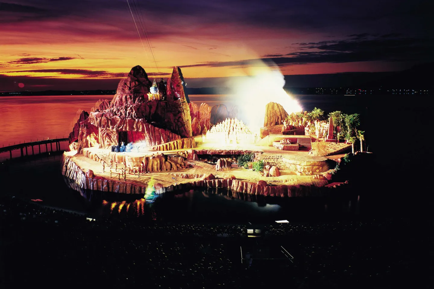 © Bregenzer Festspiele Szene aus "Die Zauberflöte", 1985 auf der Seebühne, Felsenlandschaft mit einzelnen Bäumen, ein größerer Berg befindet sich auf der linken Seite des Bühnenbilds, die Darstellenden sind mittig aufgereiht und hell angeleuchtet