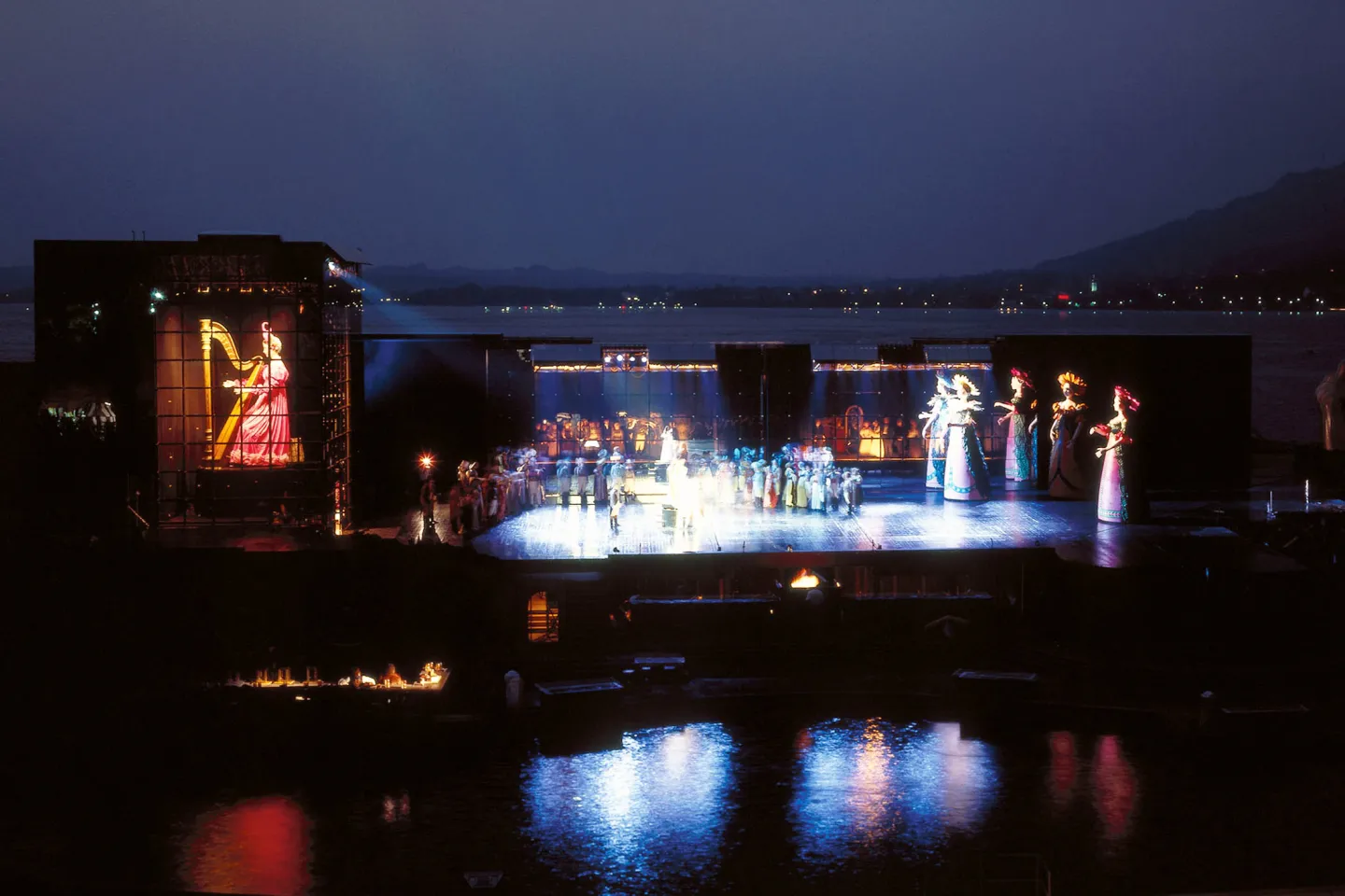 © Bregenzer Festspiele Szene aus "Hoffmanns Erzählungen", 1988 auf der Seebühne, rechts befinden sich mehrere übergroße Puppen in Blumenkostümen, links ist ein übergroßer Bildschirm mit einer Harfenspielerin, in der Mitte viele kostümierte Darstellende