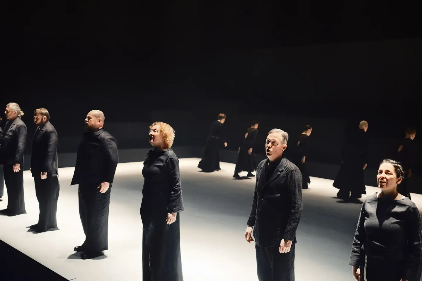 © Justus Hirvi Mehrere Personen in schwarzer Kleidung stehen in einer Reihe auf einer Bühne mit dunklem Hintergrund und hellem Boden, sie blicken nach vorne und wirken konzentriert.
