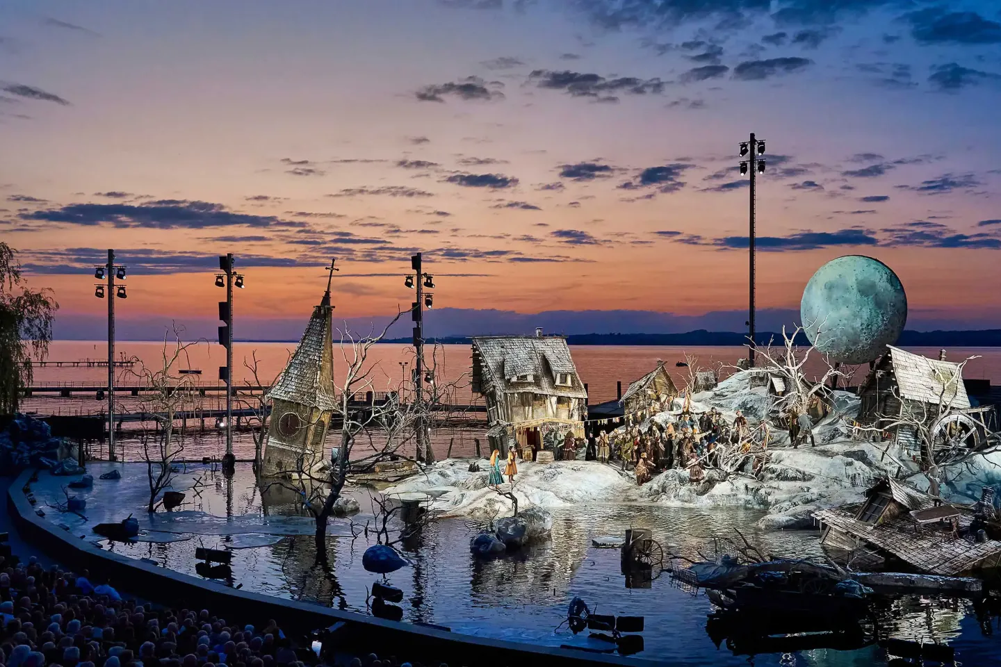© Bregenzer Festspiele / Anja Köhler Seebühne 2024/25 "Der Freischütz: Ein winterliches Dorf, überflutet, es ragt ein Kirchturm aus dem Wasser, windschiefe Häuser, über dem Schneehügel schwebt ein Mond. Im Hintergrund geht die Sonne unter, der Himmel ist rot und blau.