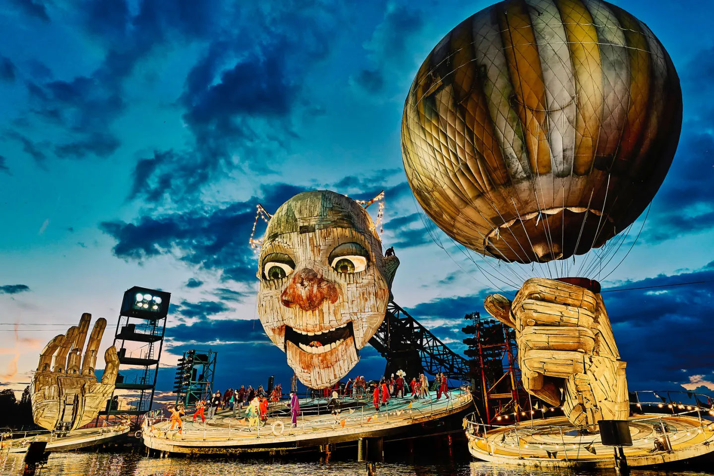 © Bregenzer Festspiele / Anja Köhler Szene aus "Rigoletto", 2021 auf der Seebühne, riesiger Kopf mit leicht geöffnetem Mund, zwei große Hände, eine Hand hält einen Ballon, mehrere kostümierte Darstellende auf der Bühnenplattform unter dem Kopf