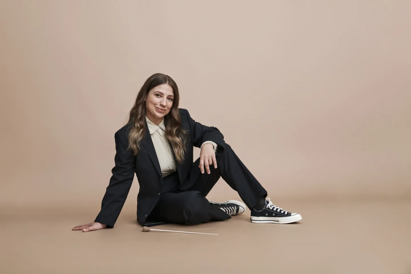 Frau sitzt auf dem Boden, trägt schwarzen Anzug, weiße Bluse und schwarze Sneakers, hält Dirigentenstab