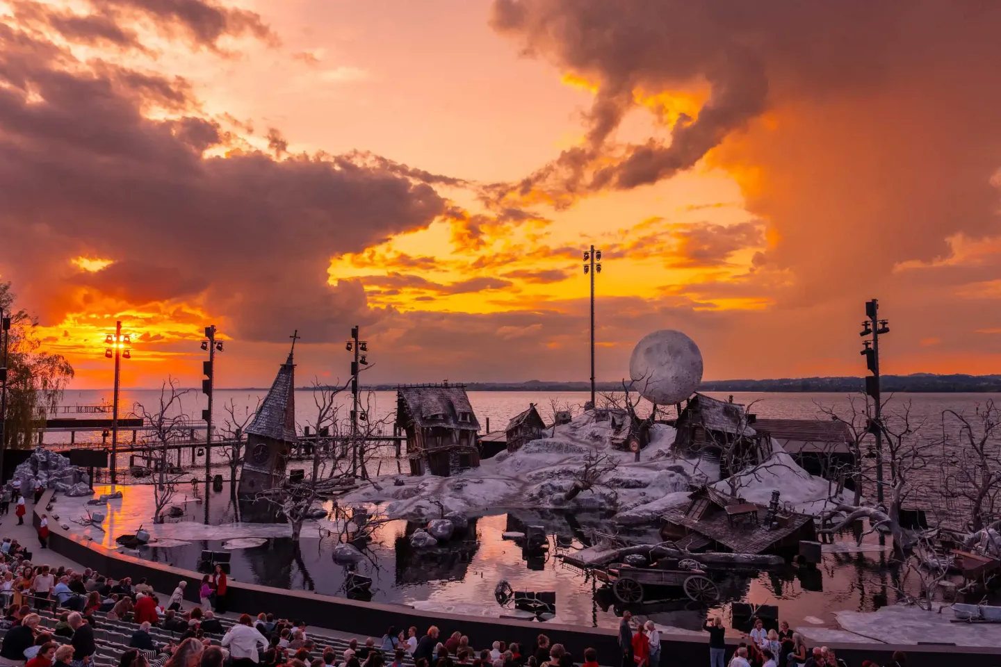 Freilichtbühne mit winterlicher Kulisse und großem Vollmond-Requisit vor einem dramatischen Sonnenuntergang am See, Publikum sitzt auf Tribünen