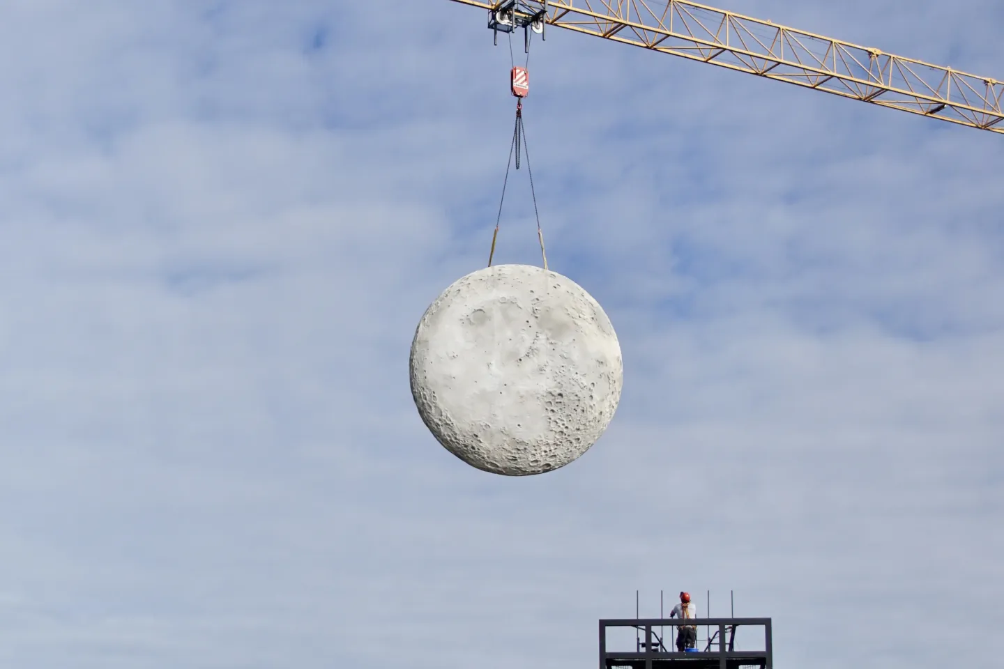 © Bregenzer Festspiele / Pia Kiker Runder, kraterübersäter Mond wird von einem Baukran an zwei Seilen über einem Gerüst mit einer Person gehoben.