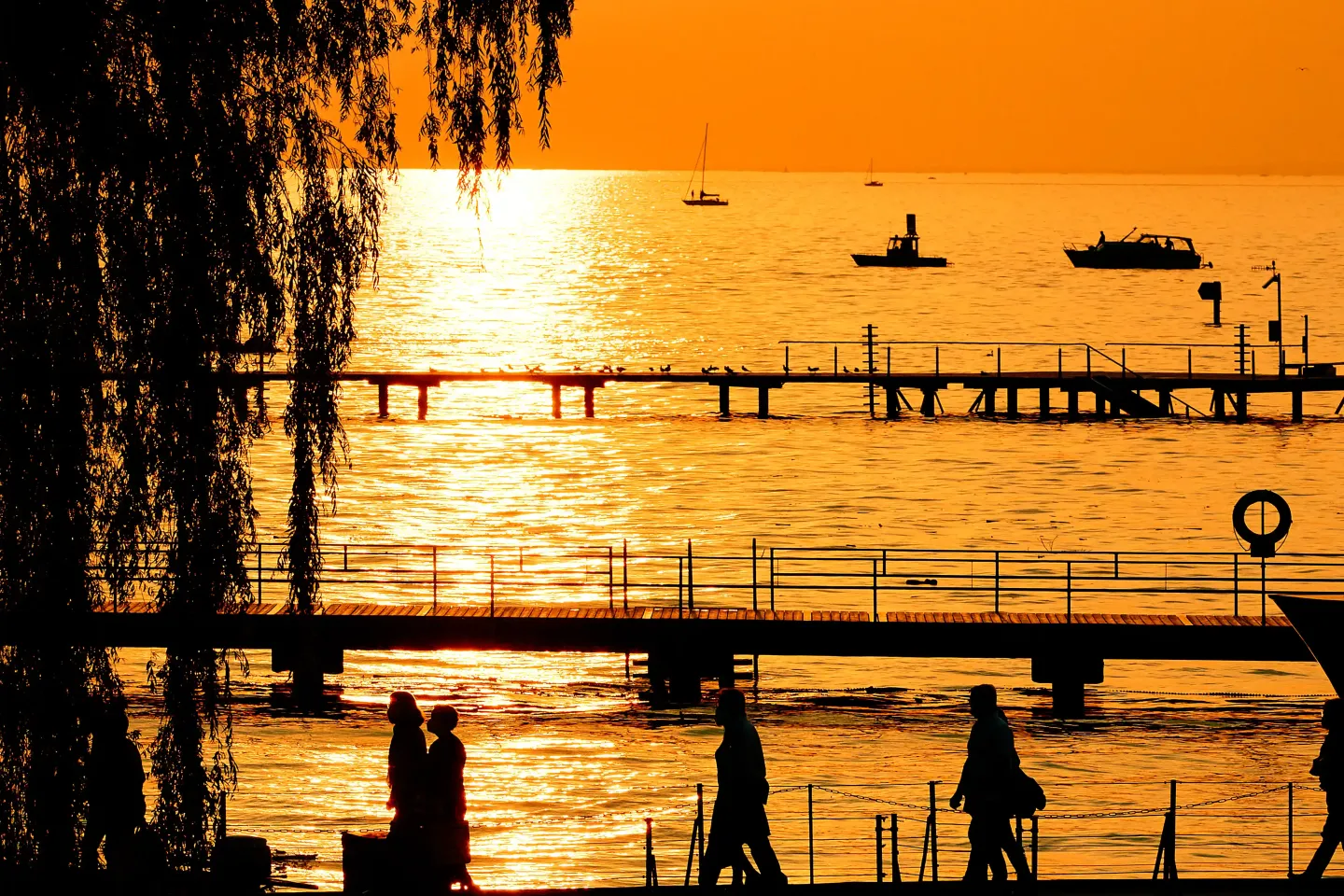 © Bregenzer Festspiele / Anja Koehler Silhouetten von Menschen auf Stegen vor einem See bei Sonnenuntergang mit orangefarbenem Himmel und mehreren Booten auf dem Wasser.