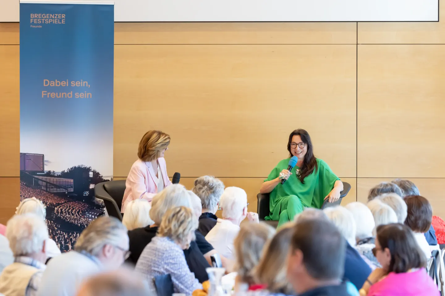 © Bregenzer Festspiele / Tatjana Schnalzger Zwei Frauen sitzen auf Stühlen vor Publikum, eine trägt ein grünes Kleid und hält ein Mikrofon, die andere ein helles Outfit. Im Hintergrund ein Banner mit Text 'Dabei sein, Freund sein'.