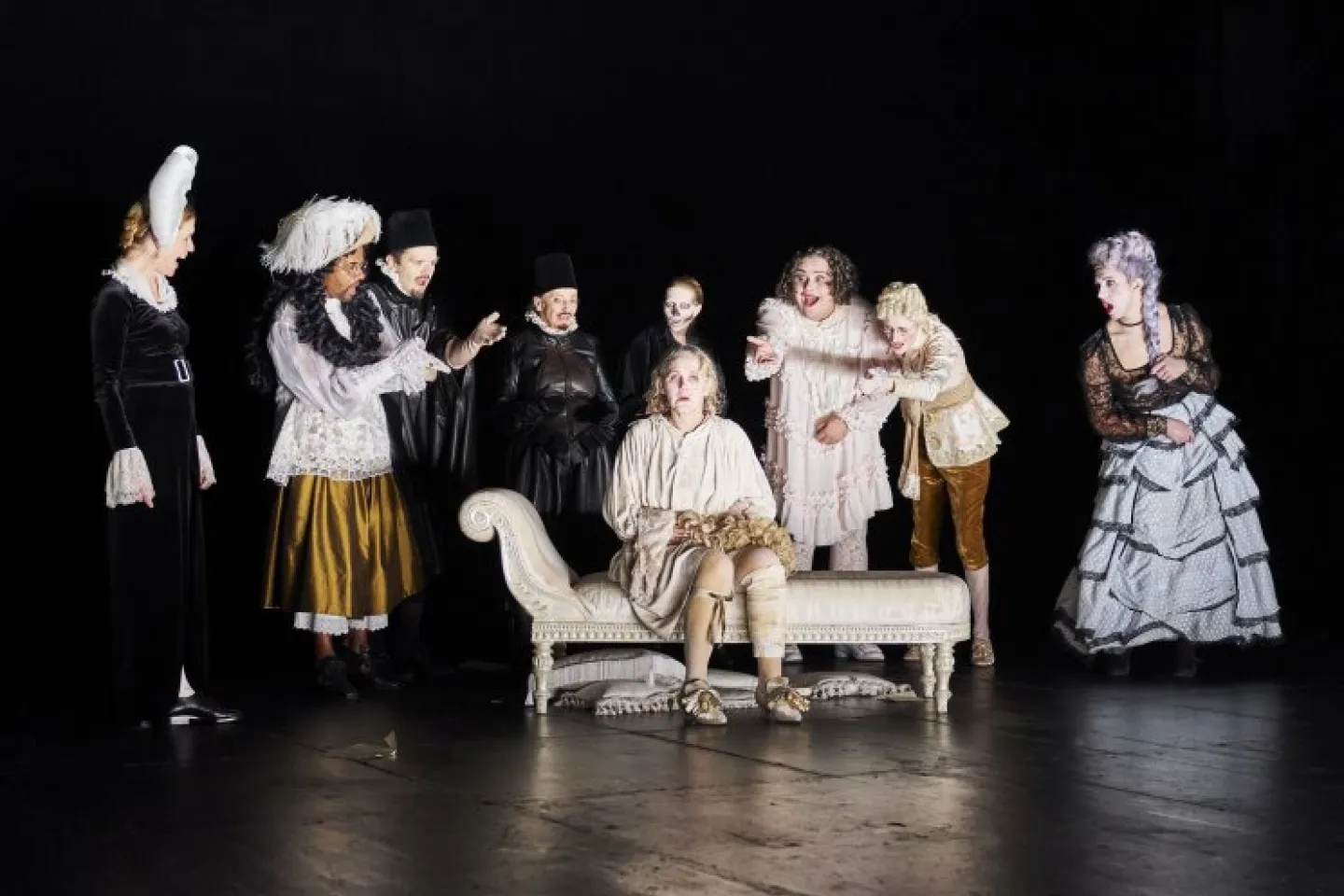 Szenenbild mit mehreren Personen in historischen Kostümen, eine Person sitzt auf einer Chaiselongue, andere stehen um sie herum auf einer dunklen Bühne.