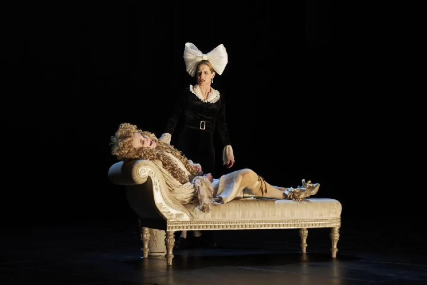 Person in schwarzem Kleid mit großer weißer Schleife steht neben liegender Person in barockem Kostüm auf einer antiken Chaiselongue.