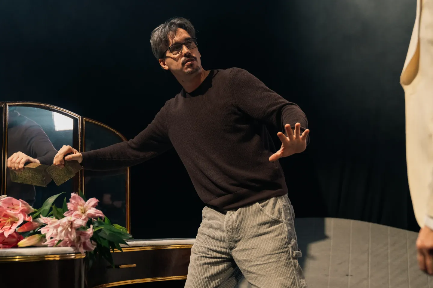 © Bregenzer Festspiele / Jonatan Langendorf Mann in schwarzem Pullover und hellen Hosen steht mit ausgestreckter Hand vor einem Spiegel mit Blumen auf dem Tisch.