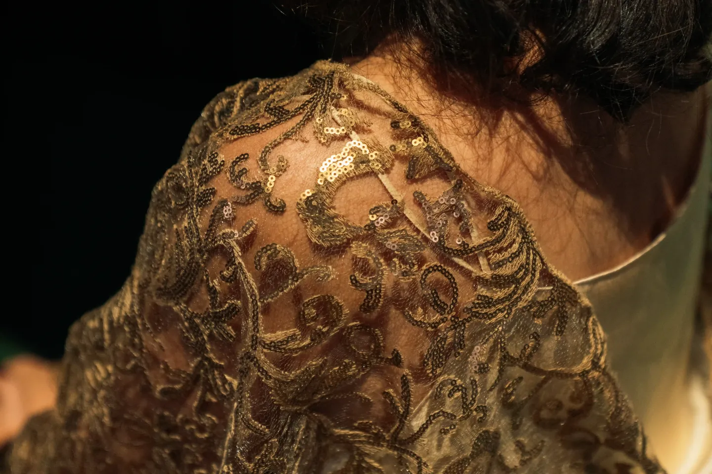 © Bregenzer Festspiele / Jonatan Langendorf Nahaufnahme eines Schulterbereichs mit goldglänzend besticktem, transparentem Stoff und dunklem Haar im Hintergrund.