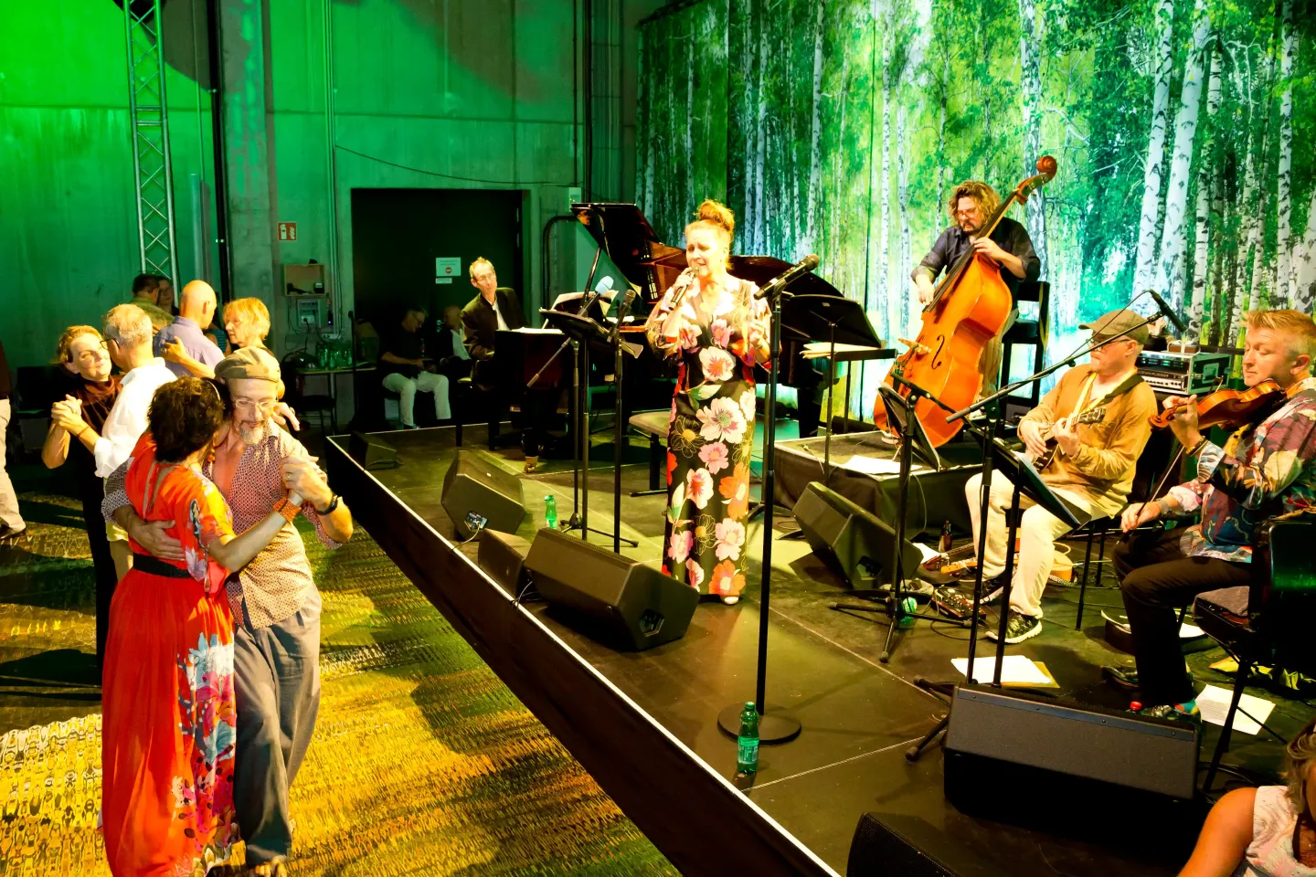 © Bregenzer Festspiele / Dietmar Mathis Frau in geblümtem Kleid singt auf Bühne mit Band, mehrere Paare tanzen davor, grüner Hintergrund mit Pflanzenmuster.