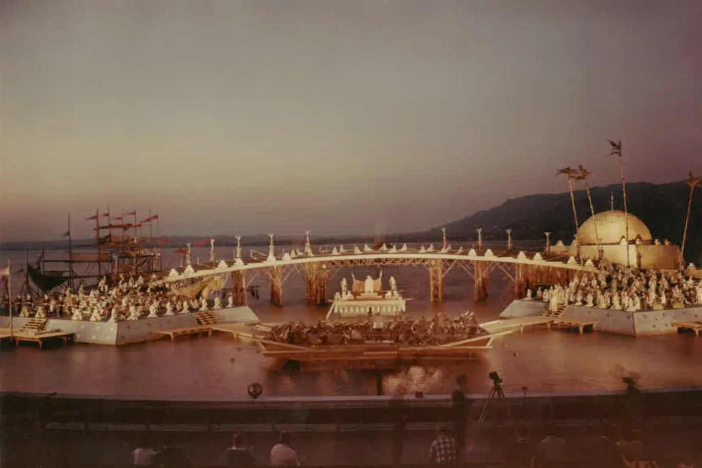 A stage set with a bridge, ships, and many figures on a body of water at dusk.