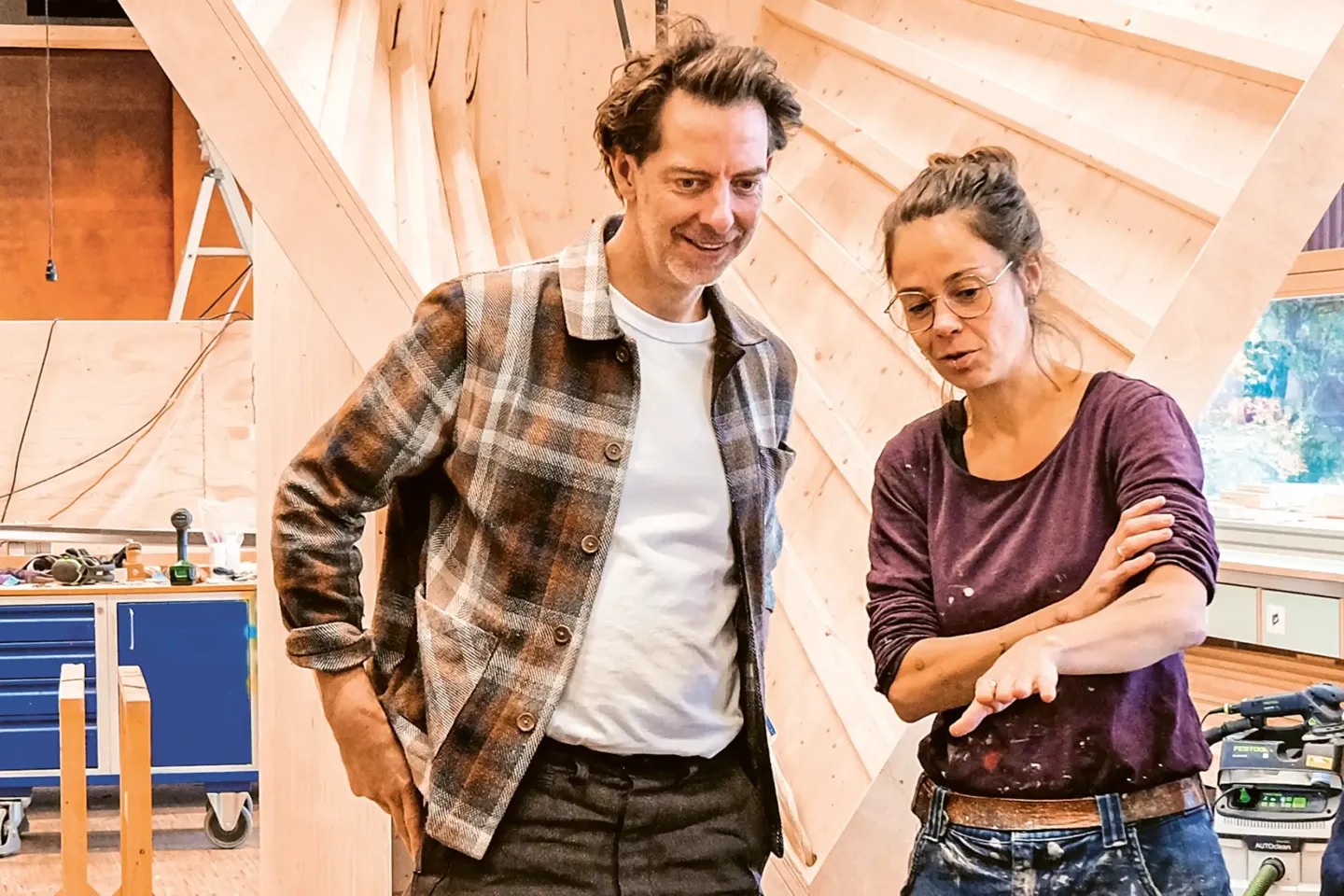 Two people are standing in a workshop with wooden structures in the background; one is wearing a plaid shirt, and the other a long-sleeved shirt with rolled-up sleeves.