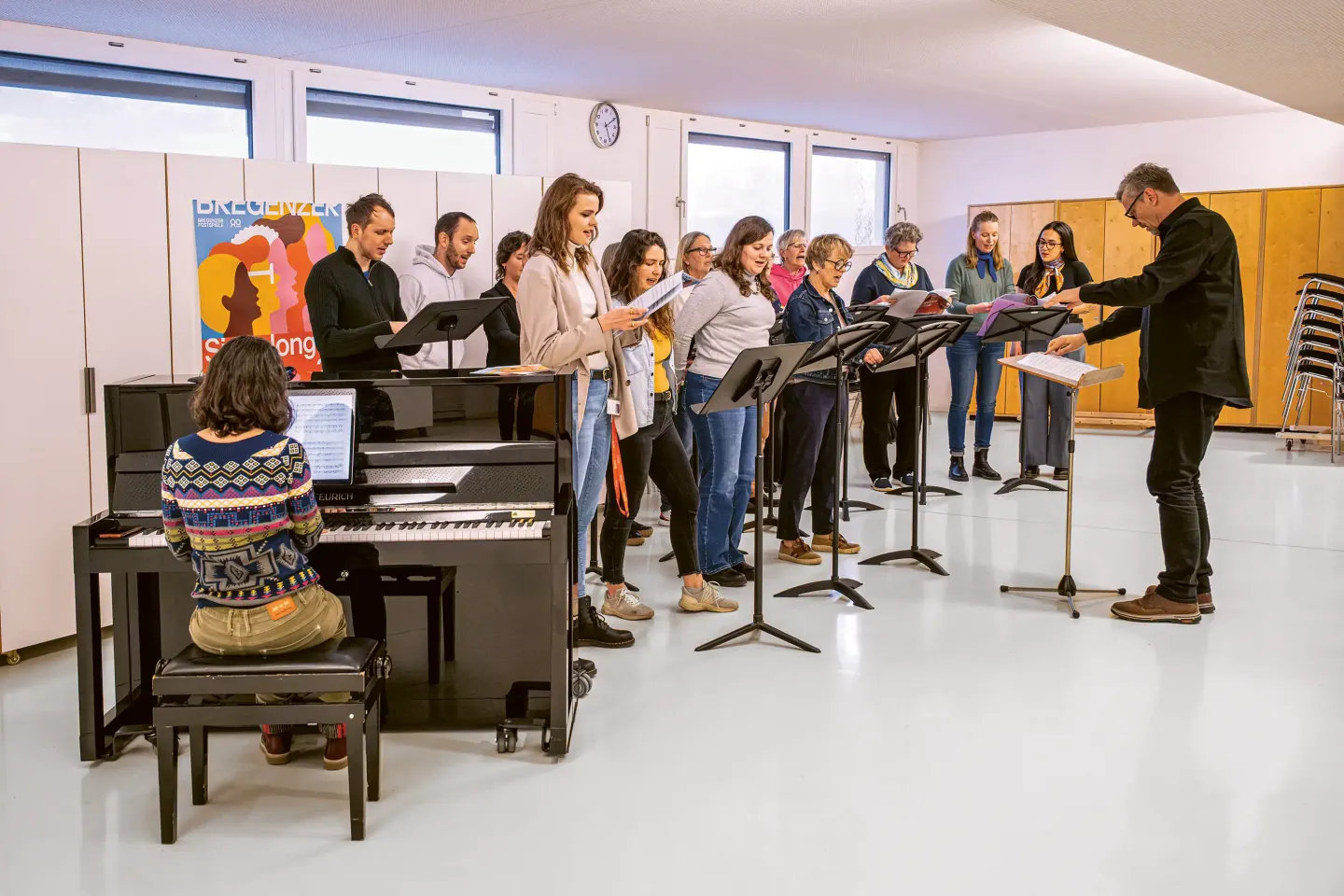 Gruppe von Menschen steht mit Notenständern in einem hellen Raum, eine Person dirigiert, eine weitere spielt Klavier