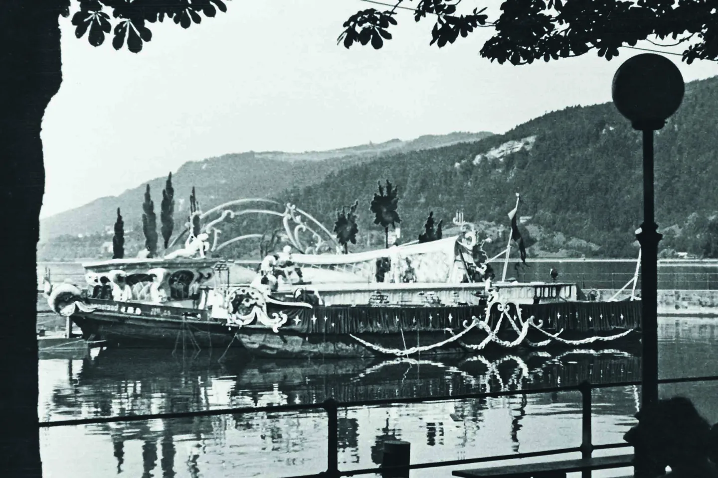 Black-and-white photo of a boat decorated with ornaments on a lake, with forested hills in the background and a tree in the foreground.