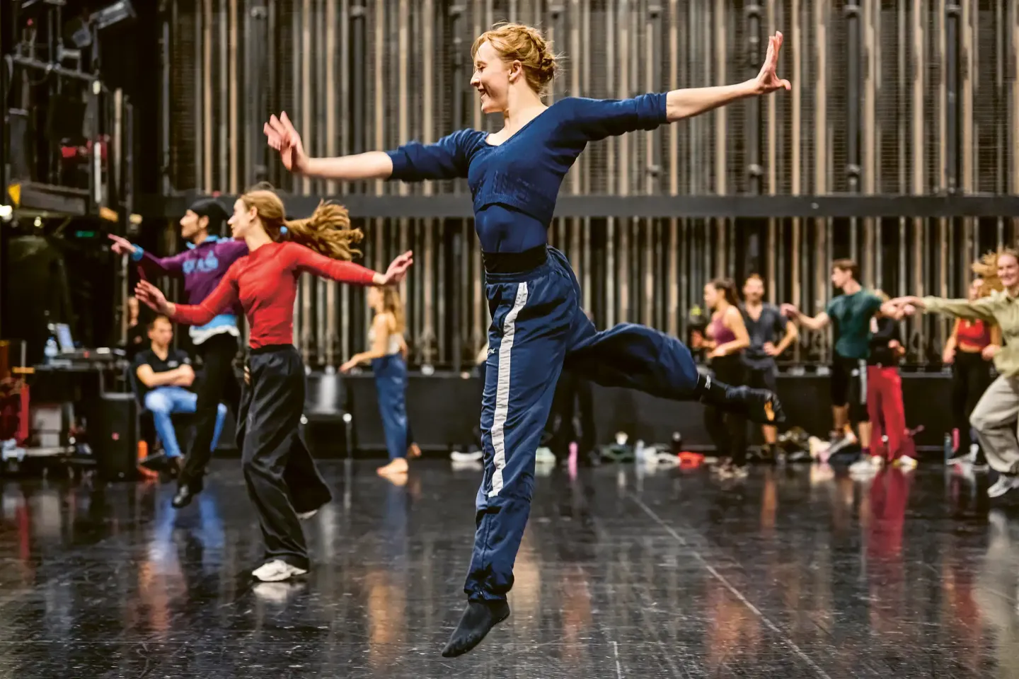 © Bregenzer Festspiele / Anja Koehler A dancer in blue training clothes is jumping with arms outstretched and one leg bent on a stage, with other dancers in the background.