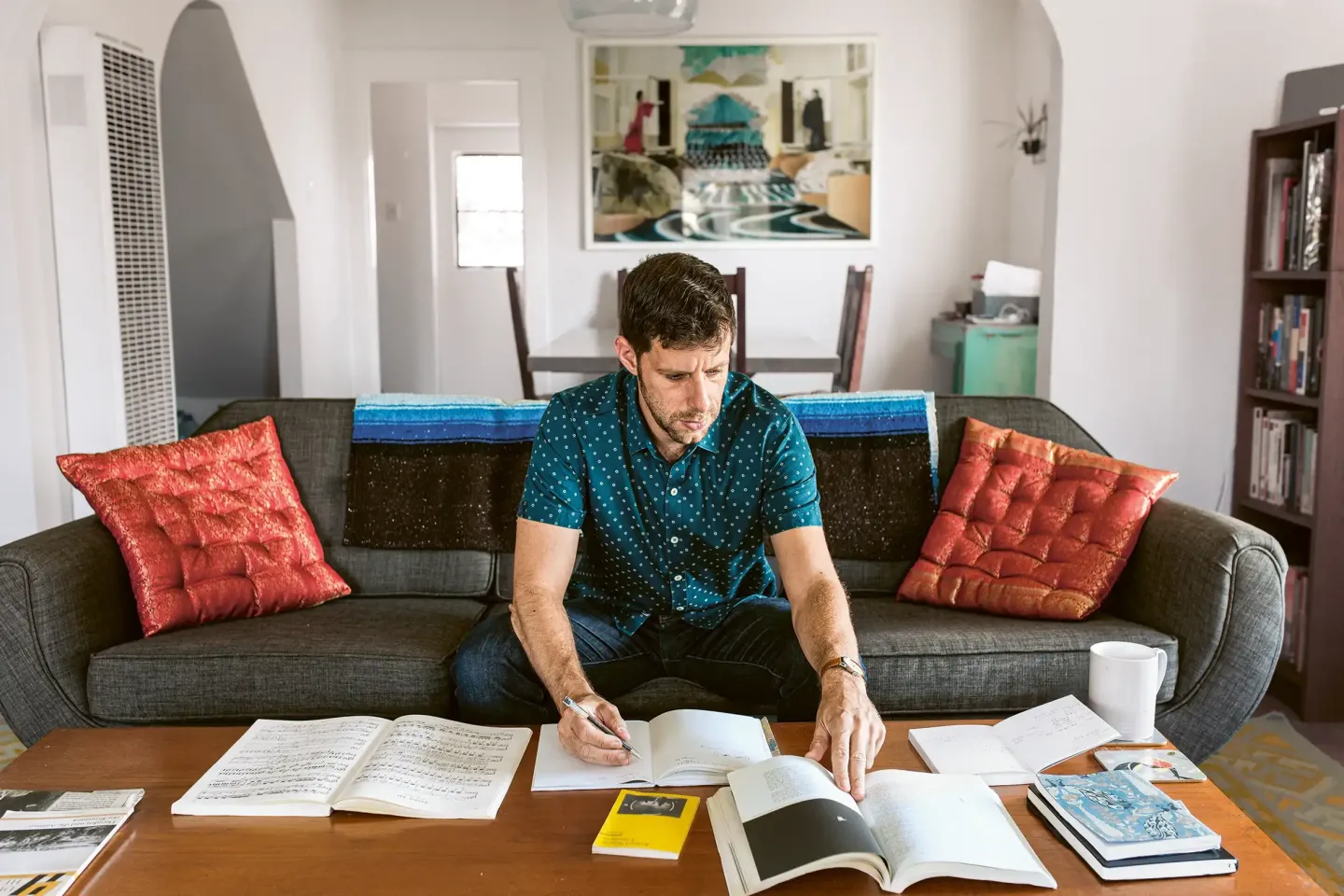 A man is sitting on a sofa, writing in a notebook in front of a table with several open books and a cup.