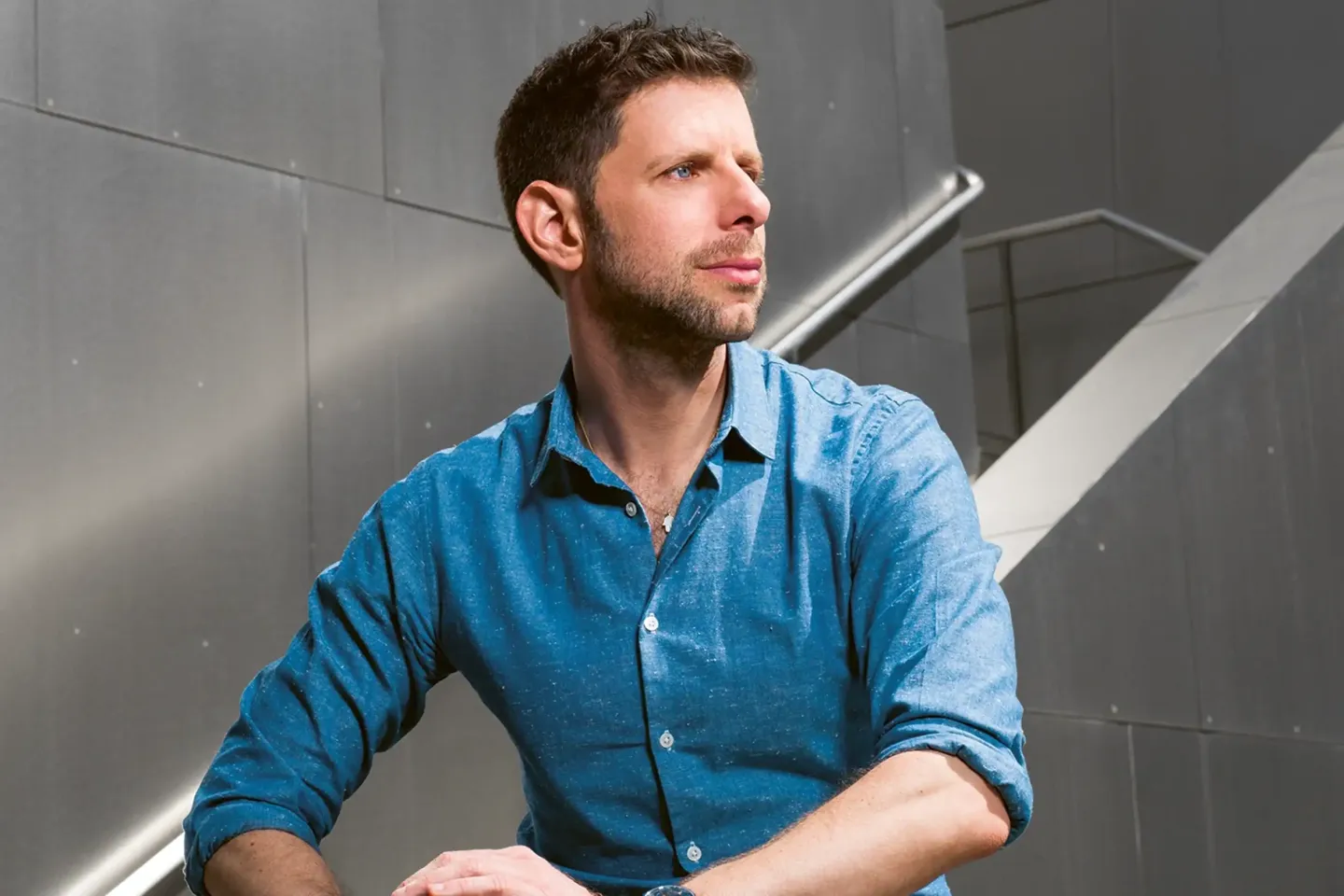 A man with a rolled-up shirt sleeve is sitting in front of a metallic wall with a staircase railing.