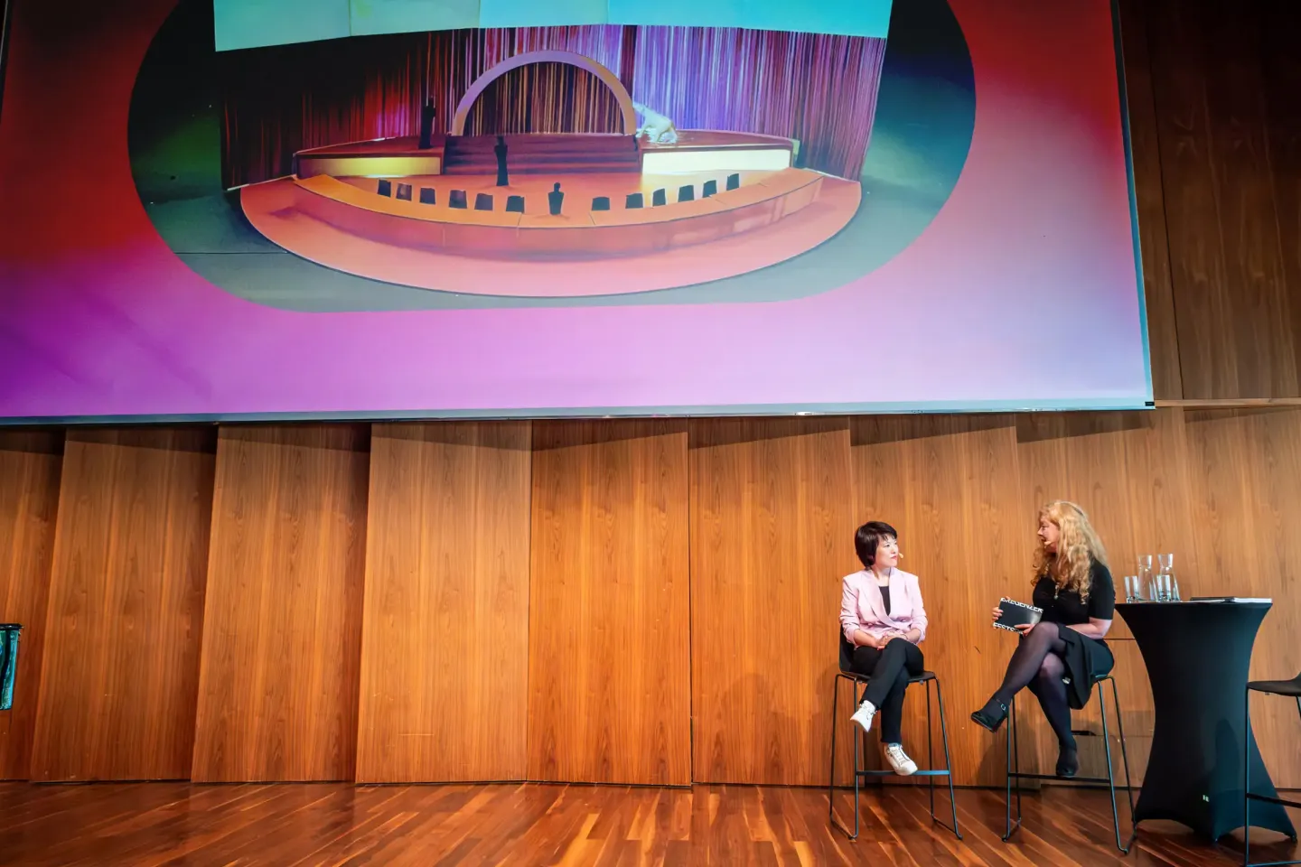 © Bregenzer Festspiele / Kristina Becker Zwei Frauen sitzen auf Stühlen auf einer Bühne vor einer Holzvertäfelung, im Hintergrund eine große Leinwand mit einer bunten Grafik
