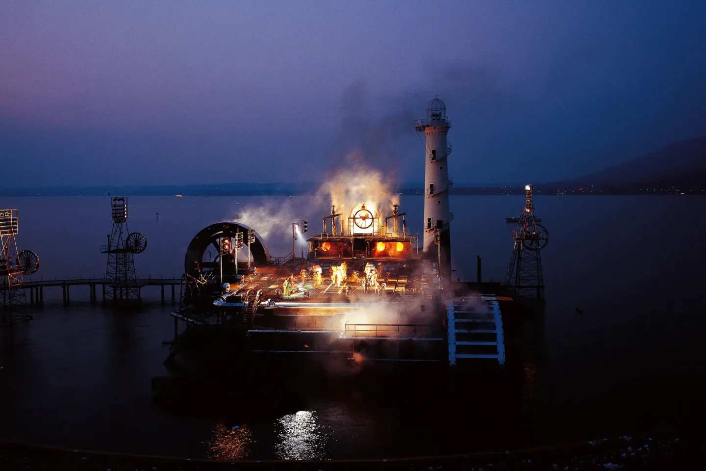 © Bregenzer Festspiele / Karl Forster Beleuchtete Bohrplattform auf dunklem Gewässer mit rauchenden und dampfenden Elementen bei Nacht