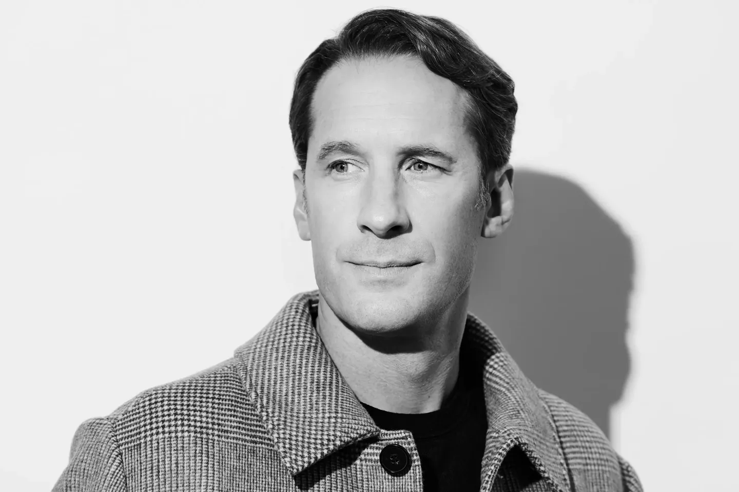 Jacques Imbrailo © Jacobus Snyman Mann mit kariertem Mantel und schwarzem Pullover vor weißem Hintergrund mit Schattenwurf