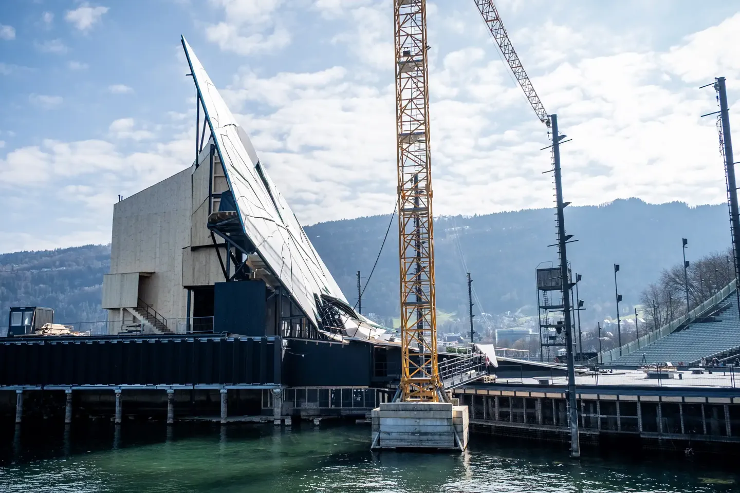 Seebühnenaufbau mit großer, schräg gestellter Wand und gelbem Baukran am Wasser vor bewaldetem Hügel.