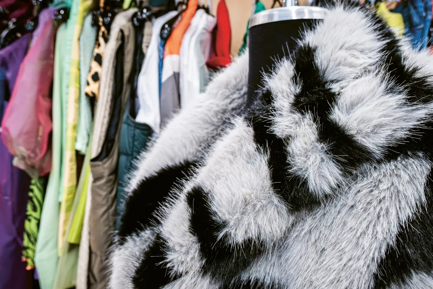 Kostüm mit schwarz-weißem Fellmuster auf Schneiderpuppe vor bunter Garderobe mit verschiedenen Kleidungsstücken.