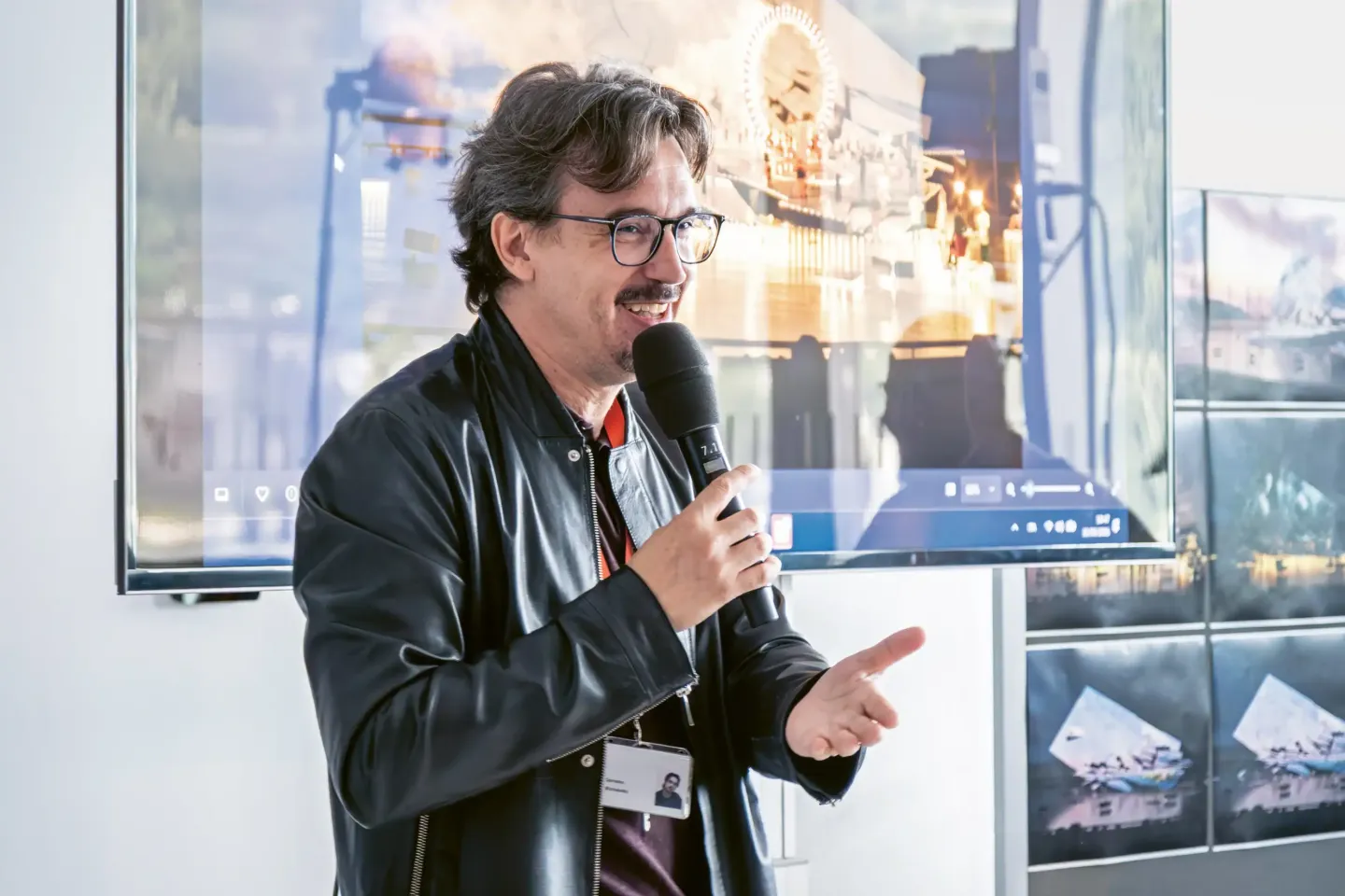 © Bregenzer Festspiele / Kristina Becker Damiano Michieletto mit Brille und schwarzer Lederjacke hält Mikrofon und spricht vor einem Bildschirm mit Abbildungen des Bühnenbilds.